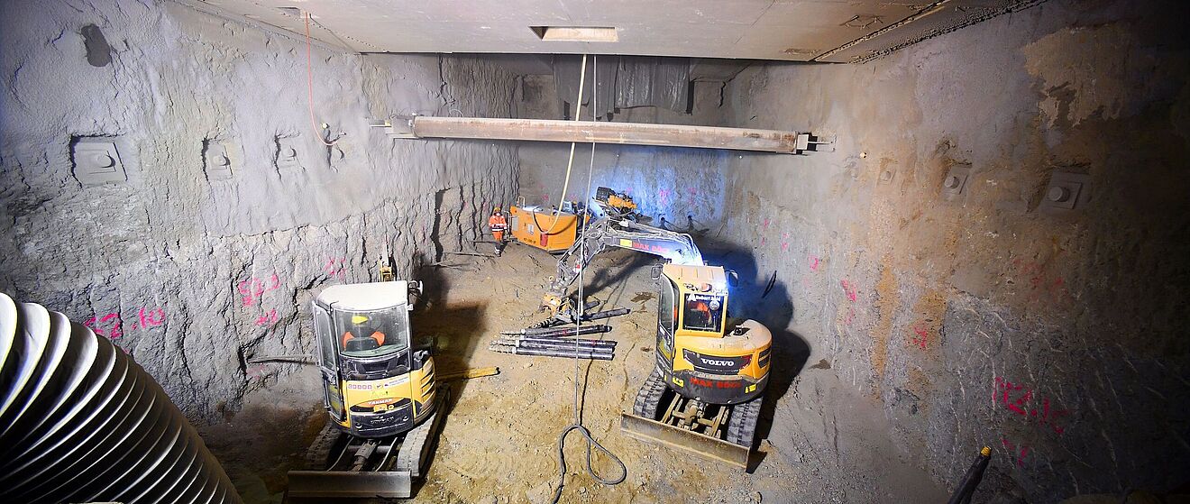 Eine unterirdische Baustelle zeigt zwei kleine, gelbe Bagger und weitere orangefarbene Maschinen. Der beengte Bereich umfasst rohe Betonwände und Erdreich.