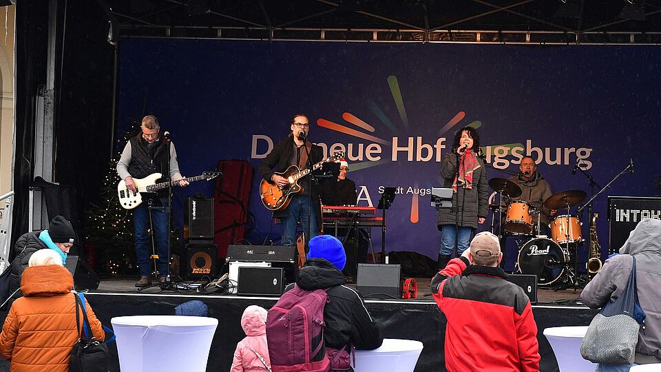 Menschen stehen an Stehtischen und blicken auf eine Bühne im Hintergrund auf der Musiker mit Instrumenten und Mikrofon stehen. Ein Klick sorgt für eine Großansicht des Bildes.